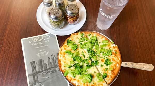 A fresh broccoli white pizza served with spice shakers and a glass of water on a wooden restaurant table.