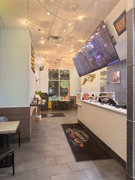 Interior of a modern restaurant featuring a textured white counter, overhead digital menu boards, warm string lighting, and VCU Rams decor.