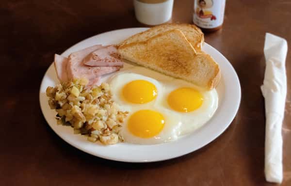 Breakfast plate with sunny-side-up eggs, toast, ham, and home potatoes.