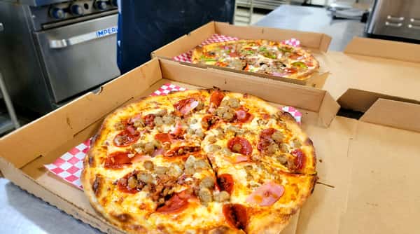 Two pizzas in cardboard boxes. The front pizza has pepperoni, sausage, ham, and bacon. The back pizza has pepperoni, sausage, peppers, onions, and mushrooms.
