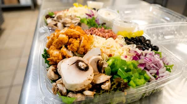 Two plastic containers of salad with fried chicken, mushrooms, shredded cheese, green peppers, red onions, bacon bits, black olives, and banana peppers.