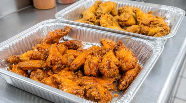 Two foil pans of chicken wings; the front pan holds orange, glazed wings, and the back pan contains dry, crispy fried wings.
