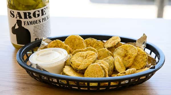 A black basket filled with breaded fried pickles and a side of white dipping sauce, next to a jar of Sarge's Famous Pickles.