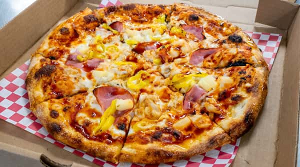 Pizza with ham, pineapple, and yellow peppers, drizzled with BBQ sauce, in a box on red and white checkered paper.