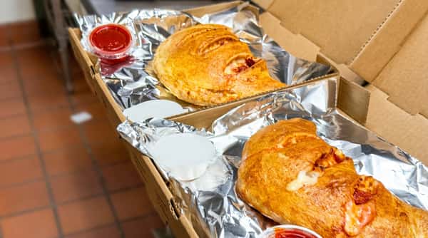 Two strombolis in a cardboard box lined with foil, with side cups of marinara and garlic sauce.
