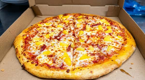 Hawaiian pizza with pineapple chunks and ham, melted mozzarella, and tomato sauce in a cardboard box.