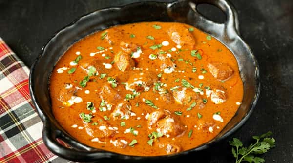 Butter chicken with a rich red-orange sauce, cream drizzles, and fresh cilantro in a black bowl.