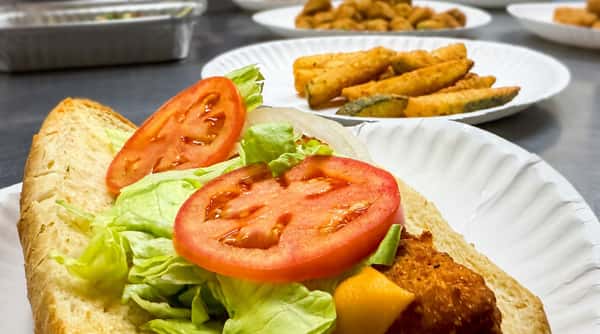 Sub sandwich with sliced tomatoes, lettuce, and fried zucchini spears on a paper plate.