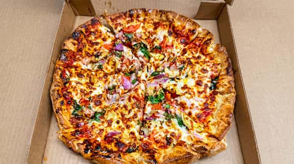 Pizza with a golden-brown crust, melted cheese, red onions, tomatoes, and green vegetables in a cardboard box.