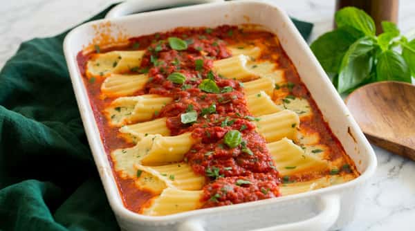 Baked manicotti in a white casserole dish, filled with ricotta and topped with red sauce, fresh parsley, and basil.