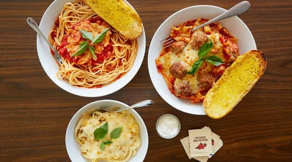 Three bowls of pasta: spaghetti with red sauce, lasagna with meatballs, and Alfredo with basil, alongside garlic bread.