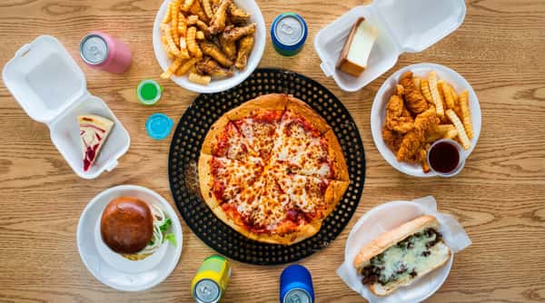 A full spread of fast food including a pizza, cheesesteak, hamburger, chicken wings, chicken tenders, french fries, cheesecake, and sodas on a wooden table.