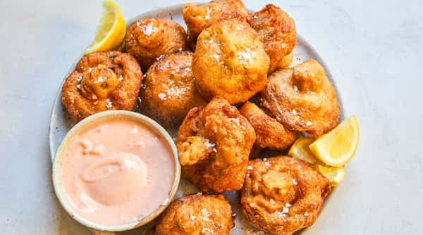 Golden brown fritters sprinkled with sea salt, served with a pink dipping sauce and lemon wedges on a light plate.