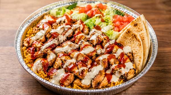 Chicken and rice platter with white sauce and hot sauce, served with shredded lettuce, diced tomatoes, and pita bread.