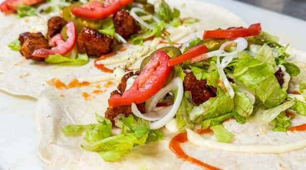 Two flatbreads topped with lettuce, sliced tomatoes, onions, jalapeños, meat cubes, white sauce, and red sauce.