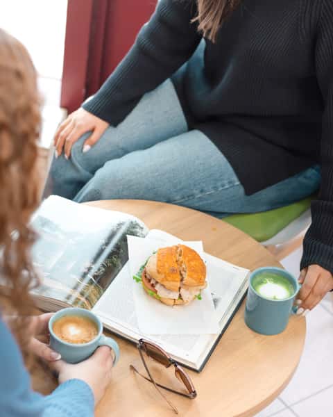 Lattes and a bagel atop a textbook, with sunglasses and two people sitting around them.