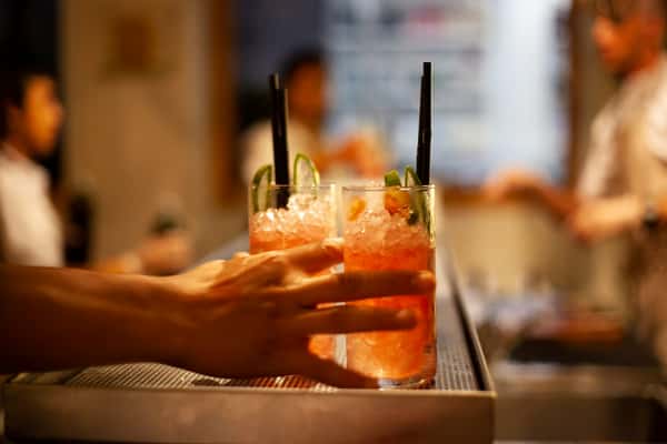 Hand reaching for two tall glasses of iced pink cocktails garnished with lime slices and served with black straws at a bar.