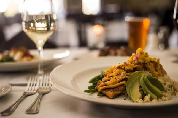 Blackened Grilled Salmon topped with avocado, mango salsa and lemon chipotle aioli served with rice pilaf on a set table with a glass of white wine.