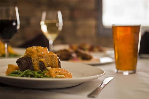 Focus of grilled beef tenderloin and sauteed shrimp with Buffalo bleu cheese cream sauce served over red skinned mashed potatoes with other plates blurred in the background. Item is accompanied by a pint of beer and a glass of white wine.