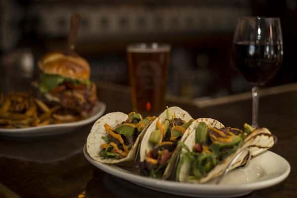 Closeup of three Steak House Tacos, Seasoned steak tips with a chipotle aioli, pico de gallo & avocado. A pint of beer and a cheeseburger are blurred in the background.