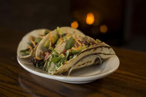 Plate of three Steak House Tacos on a wooden bar top.