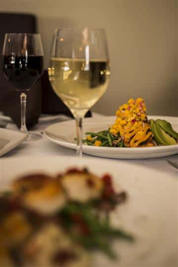 Distant view of the Blackened Grilled Salmon topped with avocado, mango salsa and lemon chipotle aioli served with rice pilaf and a glass of white and red wine.