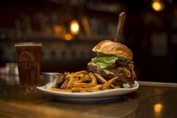 Cheese burger with a steak knife sticking in the top, topped with lettuce, tomato, caramelized onions and a side of french fries with ketchup. Served on the side is a pint of beer.