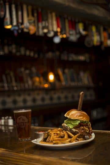 Cheese burger with a steak knife sticking in the top, topped with lettuce, tomato, caramelized onions and a side of french fries with ketchup. Served on the side is a pint of beer.