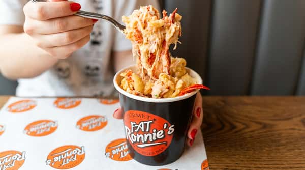 Lobster mac and cheese in a black cup from "Fat Ronnie's Burger Bar," with a fork lifting a creamy bite.