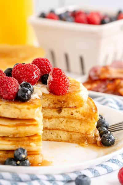 Stack of fluffy pancakes topped with fresh raspberries and blueberries, drizzled with syrup, served with extra berries and crispy bacon in the background.