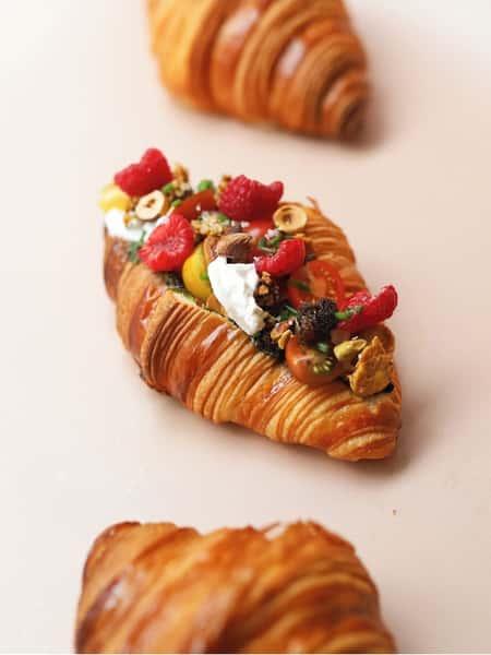 Flaky croissants filled with ricotta, cherry tomatoes, raspberries, nuts, and fresh herbs on light background