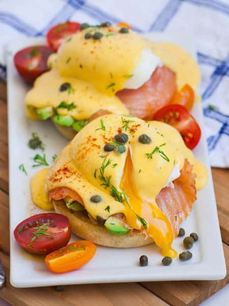 Eggs Benedict with smoked salmon and avocado on English muffins, topped with poached eggs, runny yolk, and hollandaise sauce, garnished with capers and fresh dill, served with halved cherry tomatoes.