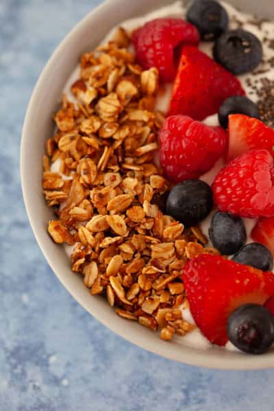 Bowl of creamy yogurt topped with granola, fresh strawberries, raspberries, and blueberries on blue surface