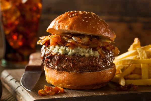 Gourmet burger with blue cheese and caramelized onions on a sesame bun, served with fries and iced drink.