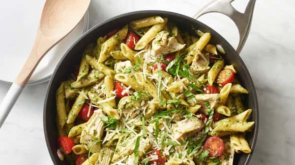Skillet of penne pasta tossed in pesto sauce with grilled chicken pieces, cherry tomatoes, and shaved Parmesan, garnished with fresh herbs.