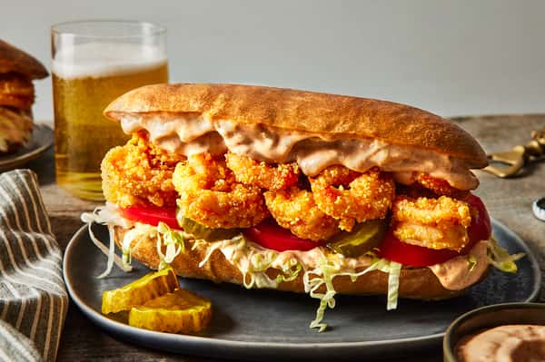 Crispy fried shrimp po' boy sandwich on a toasted roll with shredded lettuce, tomato slices, pickles, and creamy sauce, served with pickle chips and a glass of beer in the background.