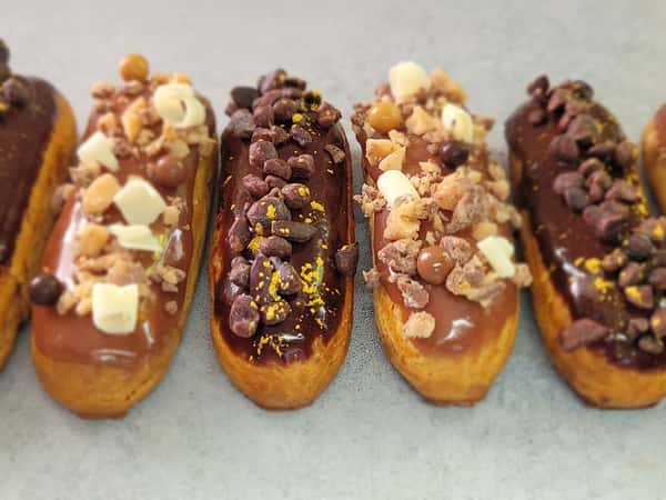 Assorted chocolate-glazed éclairs topped with various garnishes like chocolate chips, caramel bits, white chocolate chunks, and crunchy nuts, arranged in a row.