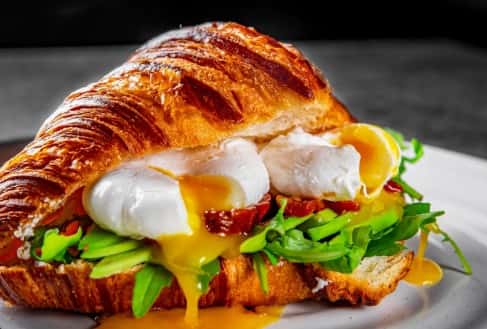 Buttery croissant sandwich filled with poached eggs, arugula, and bacon, topped with runny yolk on white plate
