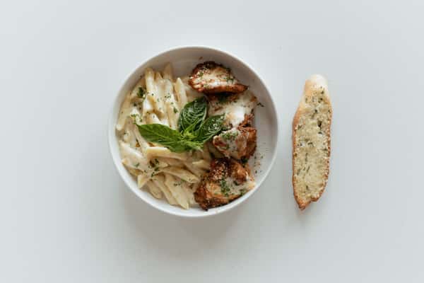 Creamy penne pasta with crispy chicken pieces, fresh basil, and grated cheese served with a slice of garlic bread