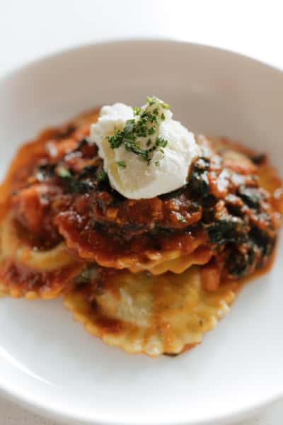 A plated stack of ravioli topped with a rich tomato-based sauce (likely meat or marinara), finished with a dollop of creamy ricotta and sprinkled herbs.
