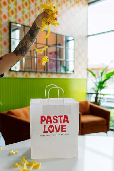 A hand tossing fresh pasta into a takeout bag labeled "PASTA LOVE," set inside a bright, modern café.