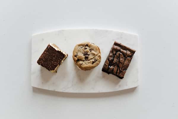 A trio of desserts on a plate: a piece of tiramisu, a chocolate chip cookie, and a brownie drizzled with chocolate.