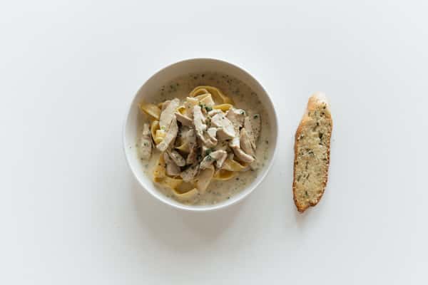 A bowl of creamy pasta (likely fettuccine Alfredo) topped with sliced grilled chicken and herbs, served with a slice of garlic bread on the side.