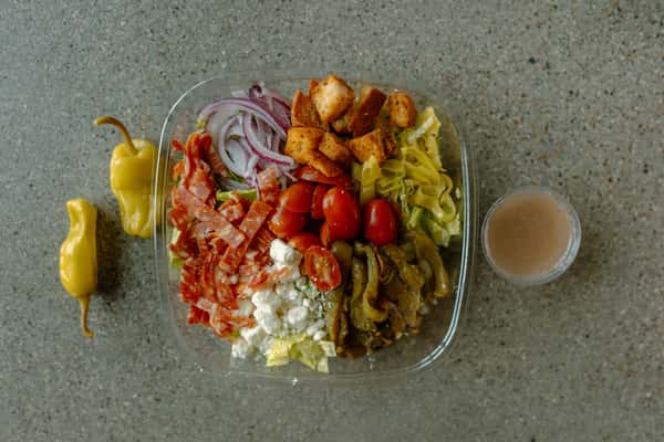 A loaded chopped salad with mixed greens, topped with sliced salami, cherry tomatoes, red onions, croutons, pickled peppers, roasted vegetables, and crumbled cheese, served with dressing on the side.