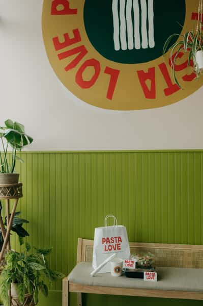 A branded "Pasta Love" display setup inside a café, featuring a takeout bag, packaged salad and pasta, and a drink arranged on a bench beneath a large wall logo, surrounded by green paneling and plants.