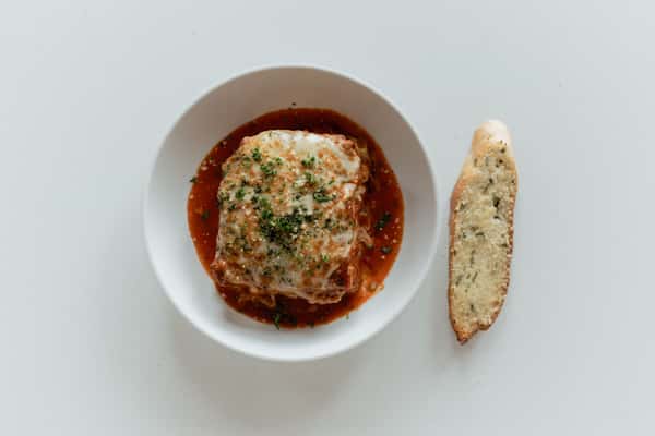 A serving of lasagna layered with meat sauce and melted cheese, topped with herbs, sitting in marinara sauce, with a slice of garlic bread on the side.