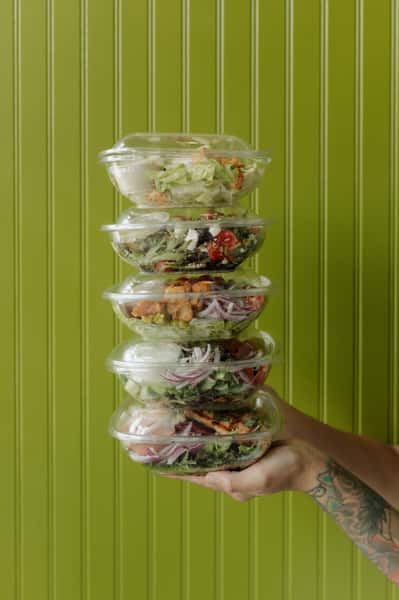 A stack of assorted fresh salads in clear plastic containers, being held in one hand against a green striped background.