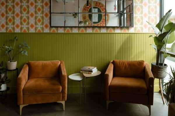 A cozy seating area in a modern café, featuring two brown upholstered chairs, a small side table with books, green paneled walls, patterned wallpaper, and several indoor plants.