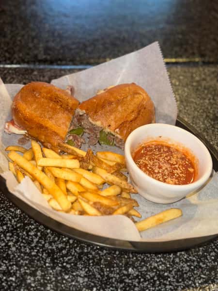 Toasted Philly cheesesteak sandwich with grilled beef, green peppers, and melted cheese, served with a side of golden French fries and a cup of marinara dipping sauce, presented on parchment paper in a black basket—classic American comfort food.
