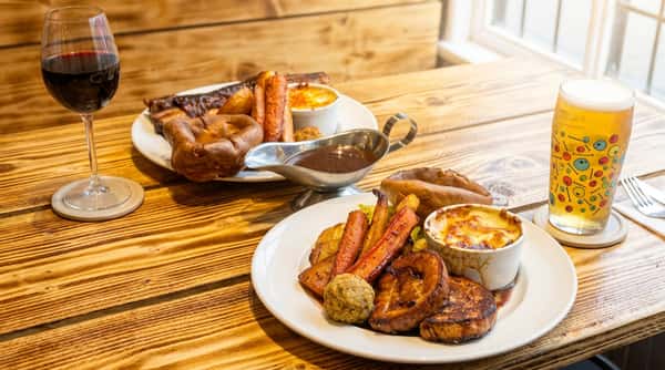 Two plates of roast dinner on a wooden table, with meat, Yorkshire puddings, roasted vegetables, and a side of gratin. A glass of red wine and a pint of beer are also visible.